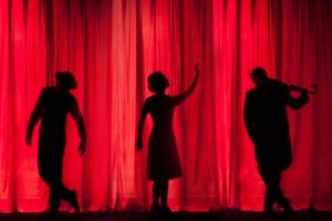 Three theatre performers shown in silhouette infront of a red curtain.
