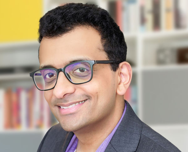 Author Aamir Hussain smiles at the camera. He has short, curly black hair and wears glasses, a dark blazer, and a light purple shirt. The background is softly blurred, showing bookshelves with colorful books and a yellow accent wall.