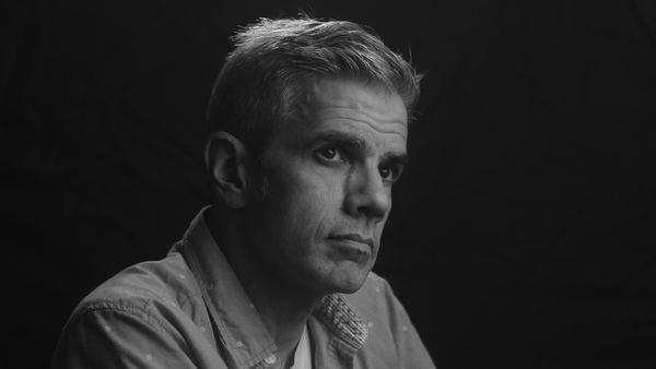 Black and white portrait photograph of author Alex Boyd. Shot in three-quarter profile, he gazes off to the left with a contemplative, serious expression. The lighting is dramatic and directional, creating strong contrast that highlights the planes of his face while leaving the right side in shadow. He wears a light-colored button-down shirt with a textured or patterned collar detail. His hair is short and slightly tousled with grey at the temples. The background is solid black, creating a classic studio portrait aesthetic with a moody, introspective tone. The composition is tightly framed from the shoulders up, emphasizing his thoughtful gaze and the sculptural quality of the lighting. The monochromatic treatment and chiaroscuro lighting give the image a timeless, literary quality appropriate for an author portrait.