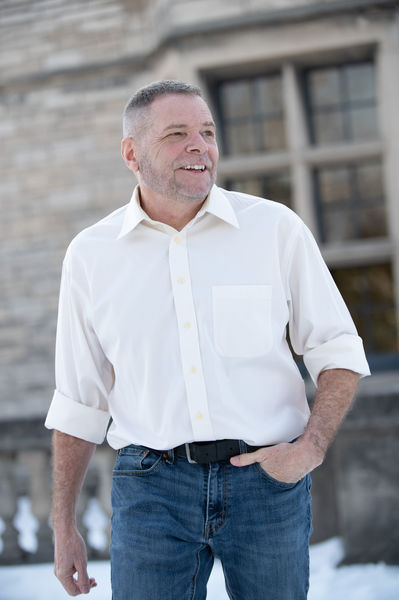 Author portrait of André Narbonne standing outdoors in natural light. He is a middle-aged man with short gray hair and a warm, genuine smile, photographed from a low angle that conveys approachability and confidence. He wears a crisp white button-down shirt with rolled three-quarter sleeves and dark blue jeans, with one hand casually in his pocket. The background shows a soft-focus stone building with large windows, suggesting a heritage or institutional setting. Bright daylight creates a clean, professional atmosphere while maintaining an informal, personable tone. The composition balances literary professionalism with accessible warmth.