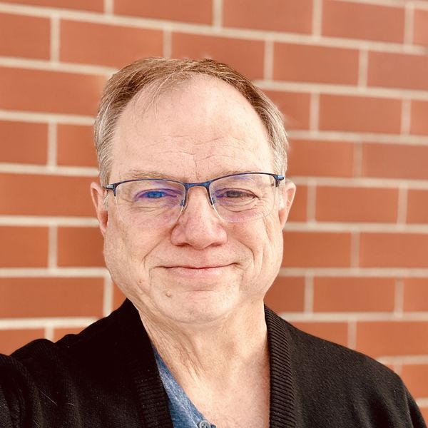Co-Editor Andy W. Taylor. A middle-aged man with short light brown hair and glasses smiles softly at the camera. He is wearing a dark cardigan over a blue shirt, standing in front of a red brick wall in bright natural light. Co-Editor Andy W. Taylor. A middle-aged man with short light brown hair and glasses smiles softly at the camera. He is wearing a dark cardigan over a blue shirt, standing in front of a red brick wall in bright natural light.