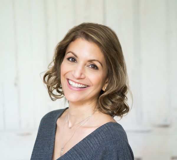 Author photo of Anita Yasuda. She is shown from the shoulders up, turned slightly toward the camera with a warm, genuine smile. She has shoulder-length brown hair with soft waves and subtle highlights, styled in a relaxed, natural way. She wears a charcoal gray off-the-shoulder sweater and delicate layered necklaces. The background is a soft-focus interior with pale, neutral tones—white or light gray walls and diffused natural light—creating a clean, airy feel. The overall mood is approachable and professional, with soft lighting that flatters without being overly stylized. The portrait conveys warmth and accessibility, fitting for a children's book author.