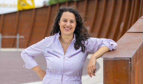 Color portrait of Anna Chatterton smiling outdoors, leaning on a rust-colored metal railing with one hand on her hip. She wears a light lavender outfit and stands in front of an abstract rust-toned structure.