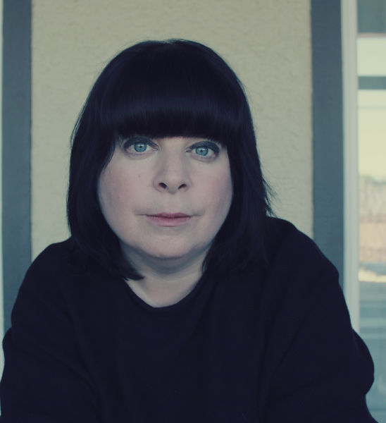 Portrait of author Anna Lazowski, shown from the chest up with straight black hair cut in a bob with blunt bangs, blue eyes, and a neutral expression. She wears a dark top and is seated indoors against a light wall with a window beside her.