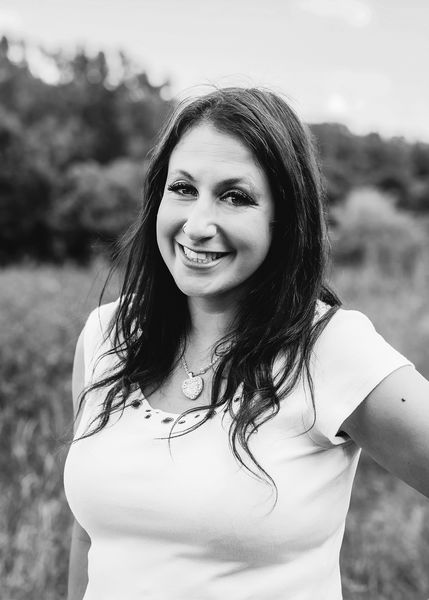 Black-and-white portrait of author Anna Baldo smiling outdoors in a grassy field with trees softly blurred in the background. She has long dark hair, wears a light short-sleeved top and a necklace with a heart-shaped pendant, and poses with one arm bent at her side. The image has a natural, relaxed feel. Black-and-white portrait of author Anna Baldo smiling outdoors in a grassy field with trees softly blurred in the background. She has long dark hair, wears a light short-sleeved top and a necklace with a heart-shaped pendant, and poses with one arm bent at her side. The image has a natural, relaxed feel.