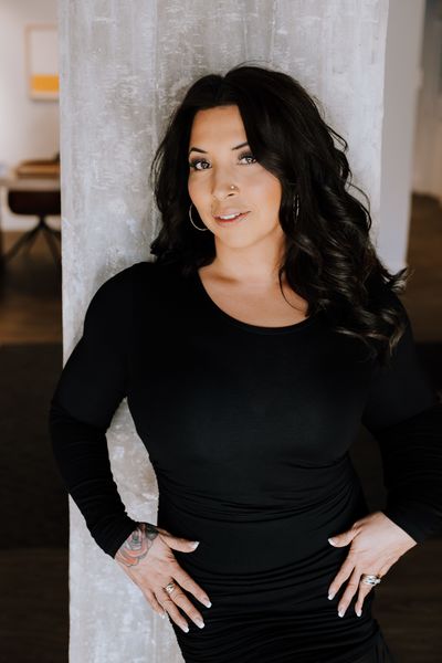 Author Arnolda Dufour Bowes stands indoors, leaning slightly against a concrete pillar. She has long, dark, wavy hair and wears a fitted black long-sleeve dress with large hoop earrings and a nose stud. Her hands rest on her hips, showing rings and a floral tattoo on her left hand. She looks directly at the camera with a confident expression.