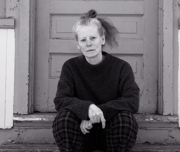 Black and white photo of author Brit Griffin, woman with light skin and curly hair tied up, sitting on worn wooden steps in front of a paneled door. She is wearing a dark sweater and plaid pants, looking directly at the camera with a neutral expression.