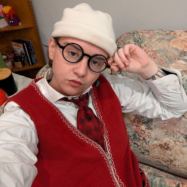 Author photo of Blaine Thornton seated on a floral couch, wearing a white knit beanie, round black glasses, a white shirt, a red vest, and a patterned tie, with one hand resting near the temple.