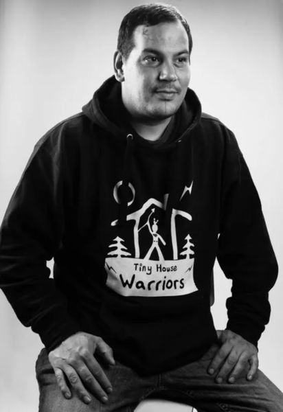 Bomgiizhik Isaac Murdoch author photo, black-and-white portrait of a man sitting and looking slightly to the side with a calm expression. He is wearing a hoodie that reads “Tiny House Warriors” with a graphic of a small house, trees, and a person raising their arms. The background is softly lit, creating a simple and focused composition.