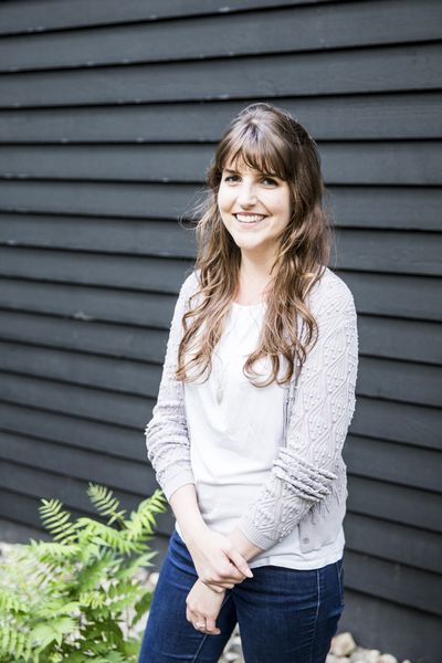 Author photo of Brittany Lane. A woman with long brown hair and bangs smiles warmly at the camera. She wears a white top with a light gray cable-knit cardigan and blue jeans, standing in front of dark gray horizontal siding with green ferns visible in the lower left corner.