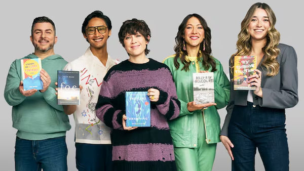Five people stand side by side against a light gray background, each holding a book in front of them. They are smiling and dressed in casual, colorful clothing. From left to right, they hold up different book covers featured in the Canada Reads competition.