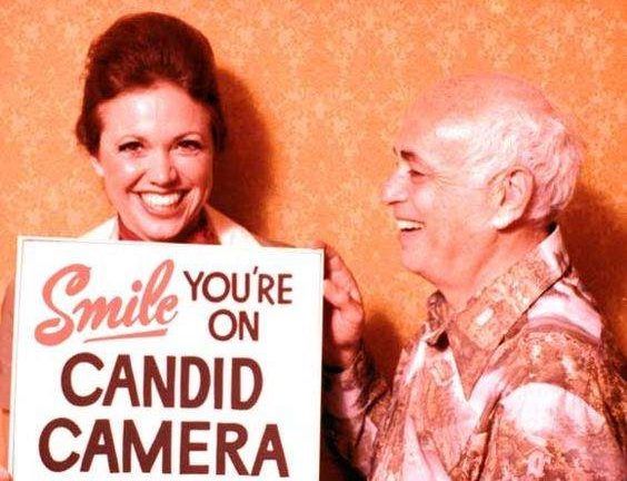 Image of a woman holding a sign that reads "Smile, You're on Candid Camera". A man stands beside her