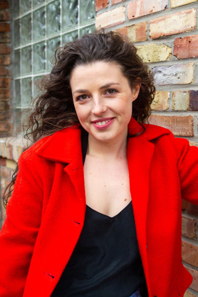 Portrait of author Charlotte Corbeil-Coleman smiling at the camera. She has long, wavy dark brown hair and is wearing a bright red coat over a black top. She stands outdoors beside a multicoloured brick wall and a glass-block window, with her hair moving slightly to one side.