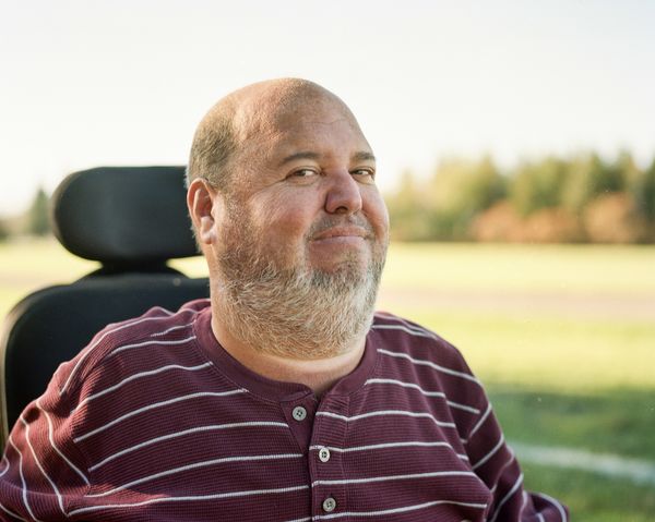 Co-Editor Matthew D. Del Papa. A man with a trimmed gray beard and short hair sits outdoors in a wheelchair, wearing a maroon and white striped henley shirt. He smiles slightly, with a grassy field and trees softly blurred in the background under bright daylight. Co-Editor Matthew D. Del Papa. A man with a trimmed gray beard and short hair sits outdoors in a wheelchair, wearing a maroon and white striped henley shirt. He smiles slightly, with a grassy field and trees softly blurred in the background under bright daylight.