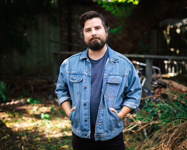 Author portrait of Curtis Campbell photographed outdoors in a wooded area with soft, dappled sunlight filtering through trees in the background. Campbell stands facing the camera with his hands casually in his jacket pockets. He has dark brown hair, a full dark beard, and wears a medium-wash blue denim jacket over a navy t-shirt. The background shows green foliage and fallen leaves on the ground, creating a natural, relaxed atmosphere. The lighting creates warm patches of sunlight on the forest floor behind him. Photo credit: Kevin Connery.