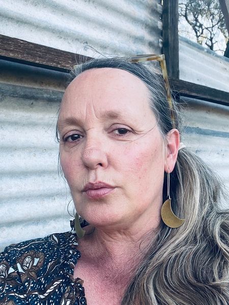 Author photo of Dani Netherclift. A woman with long, light brown hair pulled to one side poses in front of a corrugated metal wall. She is wearing large crescent-shaped earrings, a patterned top, and sunglasses resting on her head. The lighting is natural, and her expression is calm and confident.