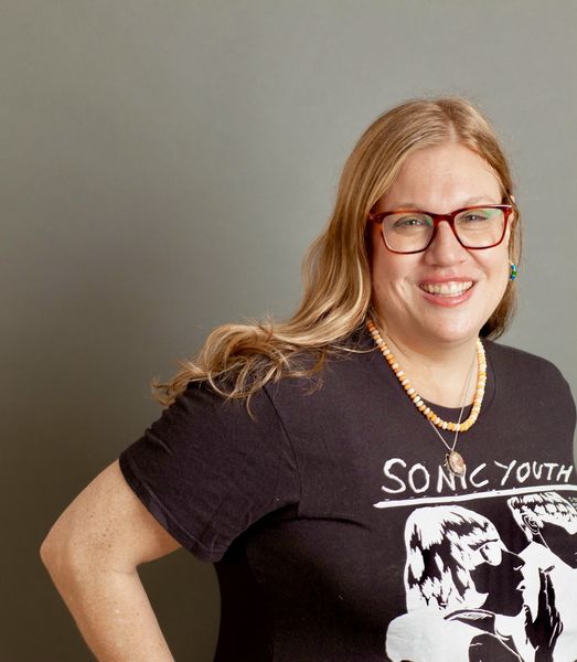Author photo of Danila Botha, featured with long light brown hair wearing red glasses, a black Sonic Youth T-shirt, and layered necklaces, smiling while standing against a plain gray background.