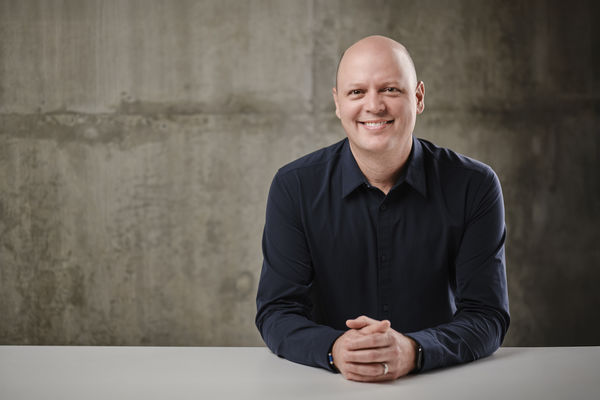 Danny Neville (Photo by Union Eleven Photographers). Portrait of a smiling man with a shaved head wearing a dark navy button-up shirt. He is seated at a white table with his hands clasped in front of him, against a textured gray concrete background.