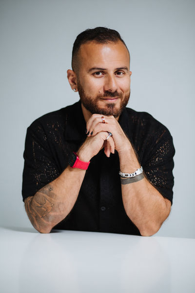 Author Danny Ramadan sits at a white table, leaning forward with his hands clasped under his chin. He wears a black shirt, a red watch, silver jewelry, and has tattoos visible on his forearm. He has a neatly trimmed beard and short hair, smiling slightly against a plain light background.