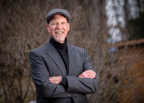 Author David Giuliano stands outdoors in front of bare tree branches, smiling warmly at the camera. He is wearing a grey flat cap, a charcoal pinstripe blazer, and a black turtleneck. His arms are folded across his chest, and he has a neatly trimmed beard and mustache. The background is softly blurred, highlighting his friendly and confident presence.