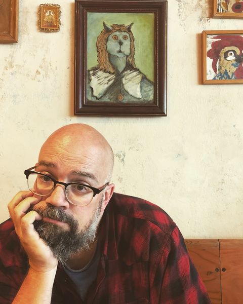Portrait of author Derek Beaulieu seated against a textured cream-colored wall. He is a bald man with a full gray beard and mustache, wearing dark-framed glasses and a red and black buffalo plaid flannel shirt over a dark t-shirt. He rests his chin thoughtfully on his hand while gazing directly at the camera with a contemplative expression. Behind him on the wall hangs a framed painting in a dark wood frame depicting a surrealist owl-headed humanoid figure with brown feathers and wide eyes, wearing what appears to be a white collar or bib. Additional framed artworks are partially visible on either side. The composition creates an artistic, literary atmosphere that complements his identity as a poet and writer. Photo credit: Kristen Beaulieu.