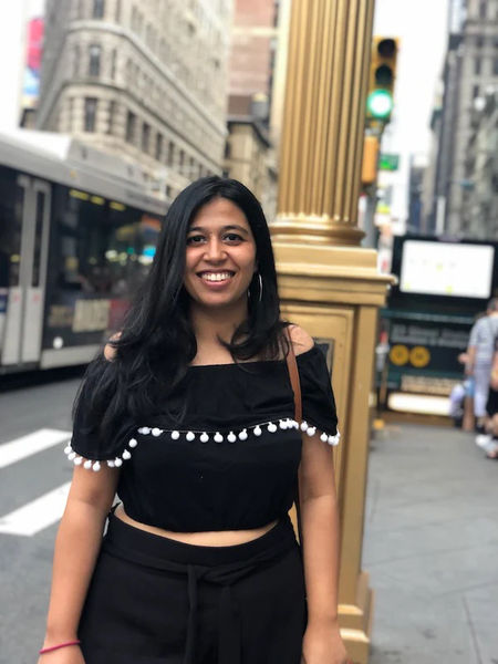 Author photo of Devika Oza taken outdoors on a city street. She stands centered in the frame, smiling warmly at the camera. She has long, dark hair worn loose and wears a black off-the-shoulder crop top with white pom-pom trim along the neckline, paired with high-waisted black pants. She accessorizes with hoop earrings and a bracelet. The background shows a busy urban intersection with classic New York City architecture—ornate building facades with detailed stonework, a tall fluted column or pillar in gold tones, traffic lights, street signs, and a city bus visible in soft focus. The lighting is natural daylight, bright and even, giving the portrait an energetic, candid feel. The overall mood is vibrant and contemporary, capturing the illustrator in a lively, real-world setting that contrasts with the more formal studio portrait style.