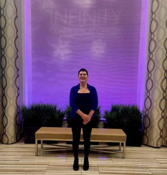 A woman stands smiling in the center of a modern, upscale interior space with a purple-lit wall behind her that reads “INFINITY CONVENTION CENTRE.” She is wearing a dark blue top and black pants with a matching blue cardigan, and stands with her hands clasped in front of her. Behind her are two tan benches and tall potted plants arranged symmetrically. The space features glossy tiled flooring and patterned columns on either side of the frame. The atmosphere is polished and professional.