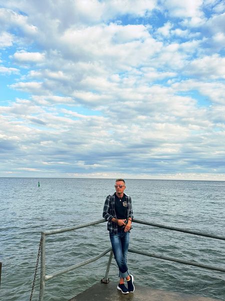 Dustin Galer stands casually on a waterfront pier, leaning against a railing with calm water behind him and a bright, cloud-filled sky overhead. He wears a plaid shirt, jeans, and sneakers. Dustin Galer stands casually on a waterfront pier, leaning against a railing with calm water behind him and a bright, cloud-filled sky overhead. He wears a plaid shirt, jeans, and sneakers.
