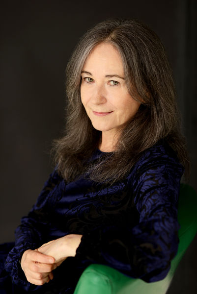 Elise Levine (Photo by Britt Olsen-Ecker). Portrait of a woman with long gray hair and fair skin, seated in a green chair against a dark background. She is wearing a dark blue, patterned top and looking toward the camera with a calm, confident expression. The lighting is soft and warm, highlighting her face and hair.