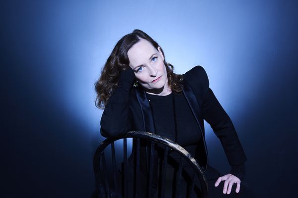 Image of the author posing on a backward wooden chair, wearing and a dark jacket over a dark shirt with one hand leant to thigh and the other up and behind her neck.