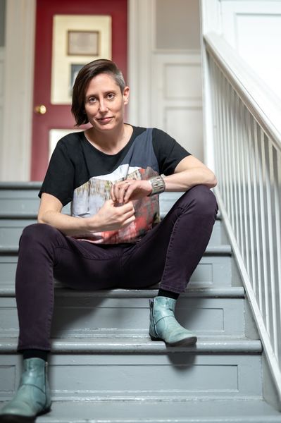 Playwright Erin Shields sits confidently on a grey staircase indoors, legs apart, leaning forward with clasped hands. She wears a black t-shirt with a bold, abstract print, dark pants, a silver cuff bracelet, and teal ankle boots. A red door with framed artwork is visible behind her, and white walls and bannisters frame the setting.