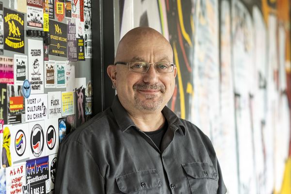 Author Francis Dupuis-Déri stands against a wall covered in colorful political stickers and graffiti art. He is bald, wearing glasses, a dark button-up shirt, and a small hoop earring. He looks directly at the camera with a slight smile.
