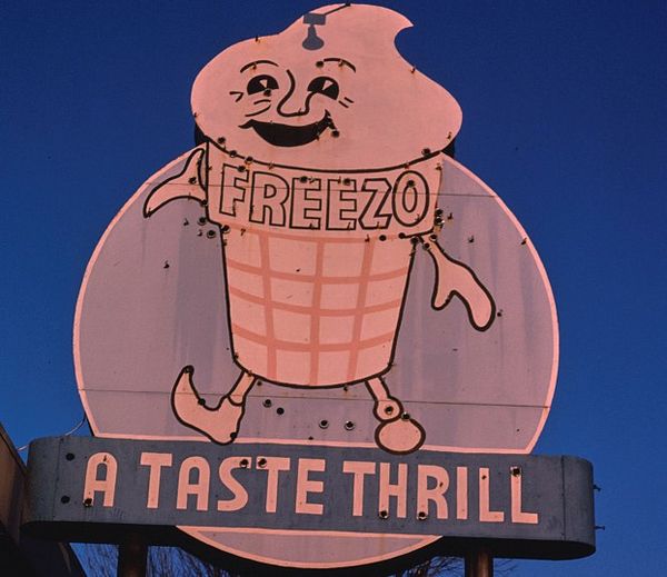 An old ice cream sign, Knoxville, Tennessee. An anthropomorphic ice cream cone smiles cheerily. He cone bears the word, FREEZO. Text beneath him on the sign reads, A TASTE THRILL.