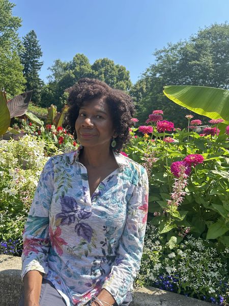 Author photo of Heather Beaumont, a woman with natural curly hair, sitting on a stone bench in a lush garden. She wears a white floral-patterned blouse with tropical leaves and flowers in shades of blue, purple, and pink, and teardrop-shaped earrings. She smiles warmly at the camera while surrounded by vibrant pink zinnias, white flowers, and large tropical foliage including banana leaves. Tall green trees and a clear blue sky are visible in the background.