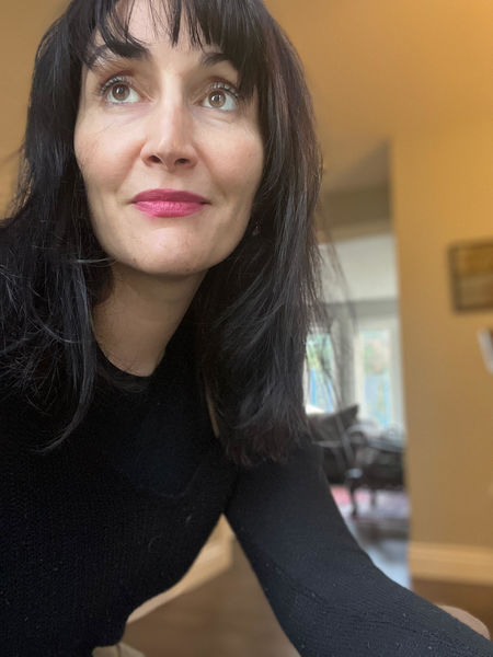 Hollay Ghadery. A woman with long dark hair and pink lipstick wearing a black top, looking slightly upward and to the side. She is indoors, with soft natural light coming from a window in the background. The setting appears to be a cozy, warmly lit room.
