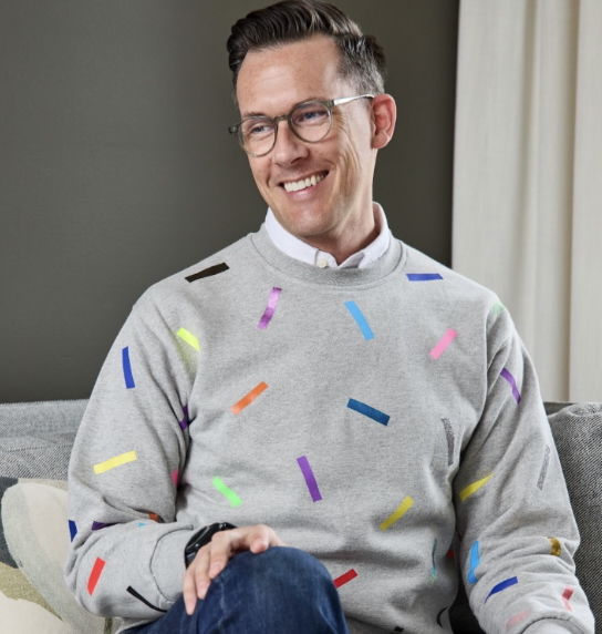 Author Ian Duncan sitting on a couch, smiling slightly to the side. He wears glasses, a light grey sweatshirt with colorful confetti-like stripes, and dark jeans. The background shows a neutral wall and curtain.