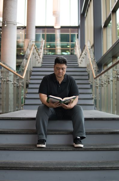 Author photo of J. Torres sitting on a wide staircase inside a modern building with tall windows and glass railings. He is wearing a black shirt and gray pants, looking down at an open book he is reading, with natural light filling the space around him.