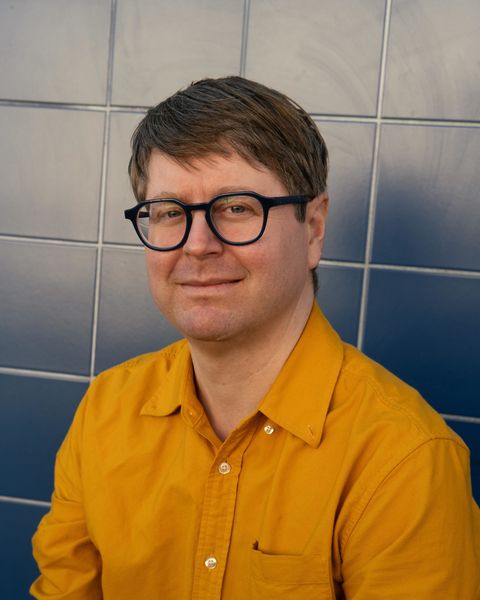 Jeff Miller (Photo by Paul Hammond). A man with short brown hair and black glasses smiling slightly while seated in front of a blue tiled wall. He is wearing a mustard yellow button-up shirt and looking directly at the camera.