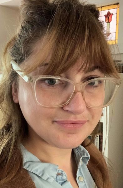 Portrait of illustrator Jennica Lounsbury, shown close up indoors with light brown hair, bangs, and large clear-framed glasses. She wears a light blue collared shirt and has a slight smile, with soft warm light and a small stained-glass window in the background.