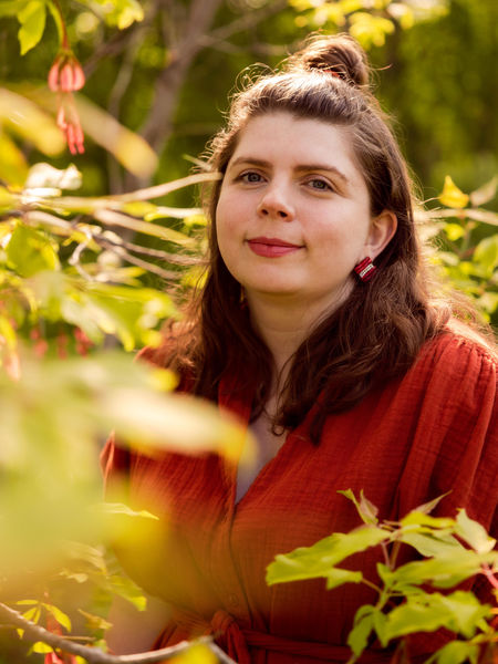 Author photo of Jess Taylor. Woman with hair in bun and long around her shoulders, wearing a woolen sweater, standing amidst a dense woodland and looking outward at reader.