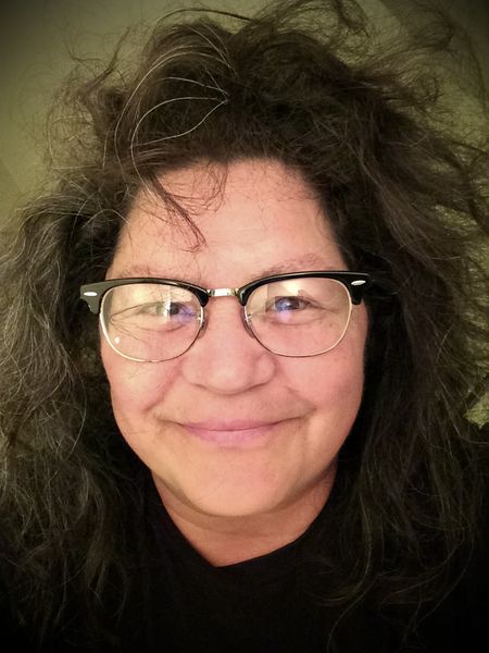 Author photo of Joanne Robertson. A person with long, dark, slightly tousled hair smiles warmly at the camera. They are wearing round glasses with dark frames and a black shirt, with a softly lit background behind them.