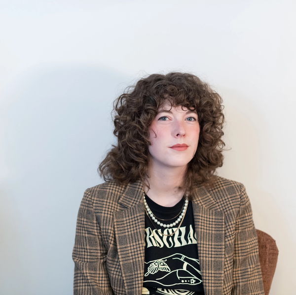 Portrait of author Joelle Kidd seated against a plain white background. She has curly brown hair and is wearing a plaid blazer over a graphic black T-shirt with light-colored text and illustrations. A strand of pearls hangs around her neck. She gazes slightly off camera with a calm, confident expression.