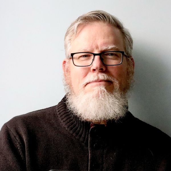 Author portrait of John Degen. He is photographed from the chest up against a plain white wall with soft natural lighting. Degen has gray hair swept back from his forehead, a full gray beard with white highlights, and wears black rectangular-framed glasses. His expression is calm and direct, with a slight upward gaze. He is dressed in a dark brown ribbed sweater over a black shirt, creating a layered, textured look. The composition is straightforward and professional, with the neutral background and soft shadow emphasizing his presence. The overall tone is approachable yet contemplative, suited for literary promotional materials.