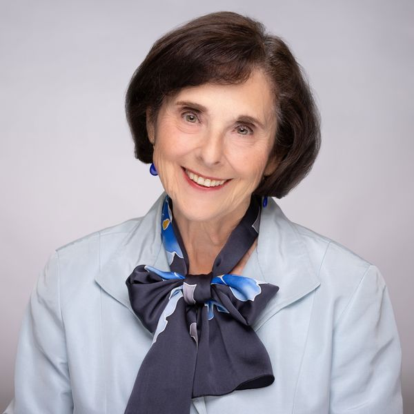 Author photo of Judith Woodsworth (Photo by Owen Egan and Joni Dufour). Portrait of an older woman with short dark brown hair, smiling warmly at the camera. She is wearing a light blue jacket and a large navy and blue patterned scarf tied in a bow around her neck, with matching blue earrings. The background is a soft gray gradient.