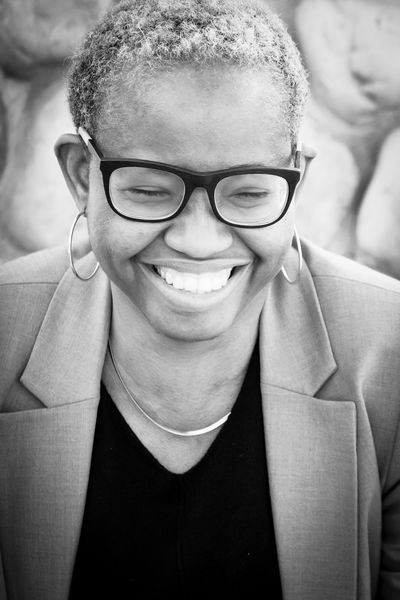 Black-and-white portrait of author Jumoke Verissimo, photographed by Cornelia Faith Photography. Verissimo is smiling warmly, wearing glasses, hoop earrings, a blazer, and a dark top, with a softly blurred stone background behind her.