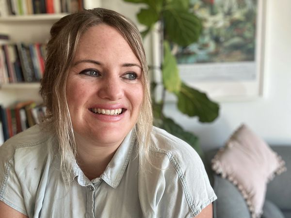 Author photo of Kasia Van Schaik. A woman with light hair pulled back and wearing a light denim shirt smiles while looking slightly to the side. She is seated in a cozy room with a bookshelf, a large green plant, and a gray couch with a pink pillow in the background.