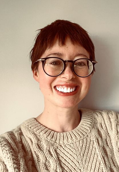 Head-and-shoulders portrait of author Katie Arthur. She smiles directly at the camera and has short reddish hair, round dark-framed glasses, and a cream cable-knit sweater. The background is plain and light-colored, keeping the focus on her face and expression.