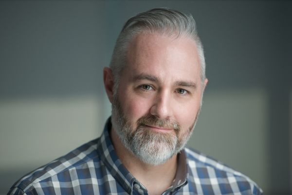 Keith Barker author photo. Portrait of a man with short gray hair and a neatly trimmed beard, wearing a blue and white plaid shirt. He is smiling softly and looking directly at the camera, with a blurred gray and beige background behind him.