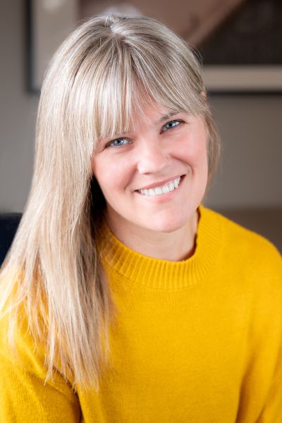 Author photo of Kelly Collier by David Gingrich. She is smiling warmly at the camera with her head slightly tilted, wearing a bright yellow knit sweater. She has long blonde hair with bangs that frame her face. The photo is taken indoors with a soft, blurred background showing neutral tones. The lighting creates a warm, professional portrait style.