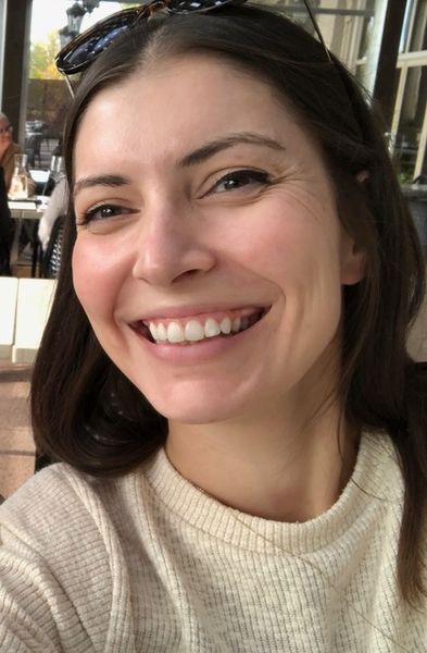 Kelsey McIver, author of St. Patrick's Day. A woman with long brown hair smiling brightly while taking a selfie outdoors. She is wearing a light beige sweater and sunglasses resting on her head, with sunlight illuminating her face.